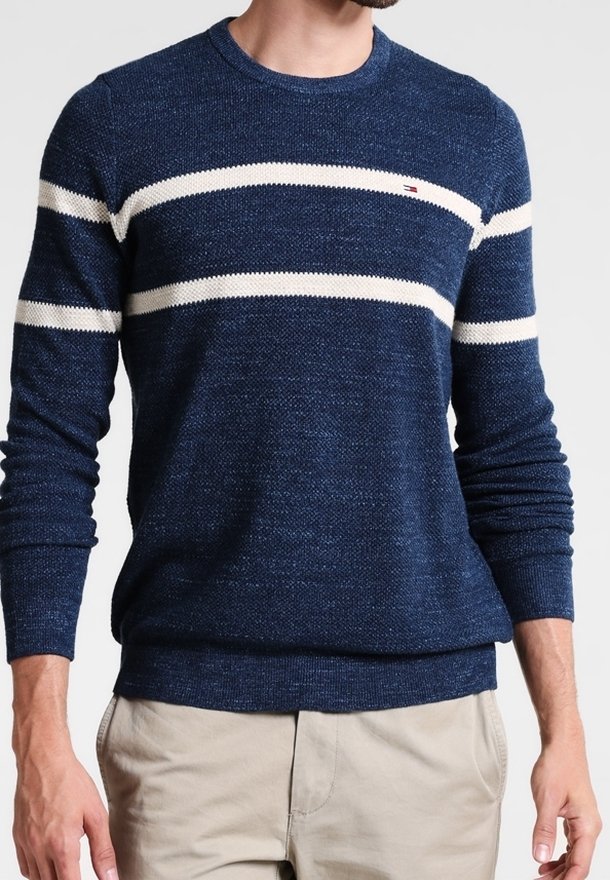 Homme portant un pull en maille bleu marine avec deux bandes blanches horizontales sur la poitrine et un pantalon beige sur fond gris.