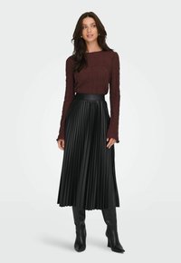 Donna che indossa un top in pizzo bordeaux a maniche lunghe, una gonna midi nera plissettata e stivali neri con tacco al ginocchio, in piedi su uno sfondo bianco.