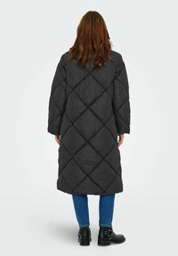 Ilgas juodas pūkinis paltas su deimantiniais siuvinėjimais, plačiomis rankovėmis ir laisvu siluetu. Dėvimas su mėlynomis džinsomis ir kulkšnies batais.