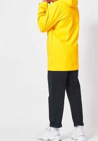 Gelbe wasserdichte Jacke mit Kapuze, kombiniert mit schwarzen schmal geschnittenen Hosen und weißen Sneakers. Die Jacke hat eine glatte Textur und ein minimalistisches Design.