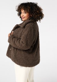 Bruine, pluizige, textuurjacket met een kraag en knoopsluiting, gecombineerd met crèmekleurige broek. De jacket heeft een losse pasvorm en een zachte stof.