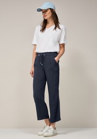 Haut blanc à col en V à manches courtes associé à un pantalon bleu marine avec cordon de serrage et des baskets blanches, complété par une casquette bleu clair.