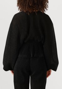 Zwarte gebreide top met een textuur, brede lange mouwen en een elastische taille. De stof heeft een subtiele glans, die een geribbeld patroon creëert.