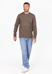 Braunes Langarmhemd aus weichem Material, kombiniert mit hellblauen Jeans. Das Hemd hat einen runden Halsausschnitt und einen lässigen Schnitt. Weiße Sneaker runden den Look ab.