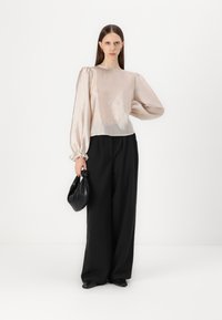 Lys rosa strukturert bluse med puffede lange ermer, kombinert med svarte vide bukser. Modellen holder en liten svart veske.