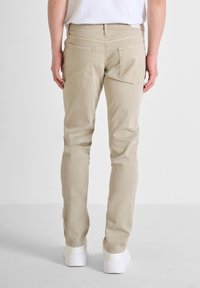Pantalones de algodón beige con corte ajustado, que cuentan con dos bolsillos traseros y un diseño minimalista. Llevados con zapatillas blancas sobre un fondo claro.