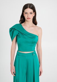 Türkisfarbenes Top mit einem Schulterträger aus glattem Stoff und einem drapierten Ärmel. Cropped-Design, minimalistische Silhouette und kombiniert mit passenden, plissierten Hosen.