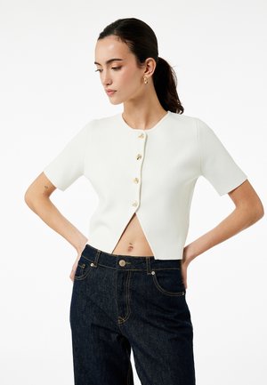 Haut blanc à manches courtes avec un col rond, fermeture à boutons sur le devant, tissu texturé et design court, associé à un jean bleu foncé.