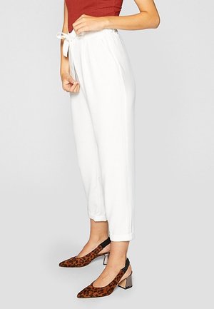 Woman wearing white drawstring pants and brown leopard print slingback heels, standing against a plain light background.