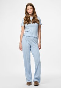 Mlada ženska z dolgimi valovitimi lasmi, oblečena v svetlo modro pleteno majico s kratkimi rokavi in ujemajoče hlače, ki stoji na beli podlagi.