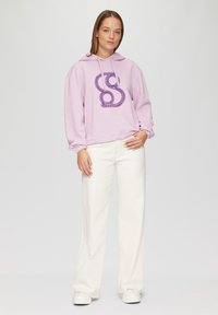 Hoodie lilla con un design grafico viola, abbinato a pantaloni bianchi a gamba larga e sneakers bianche. Presenta una vestibilità comoda e un cappuccio.