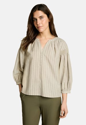 Frau mit langen braunen Haaren, die eine beige Bluse mit vertikalen Streifen und 3 Knöpfen sowie olivgrüne Hosen trägt, steht mit einer Hand in der Tasche.