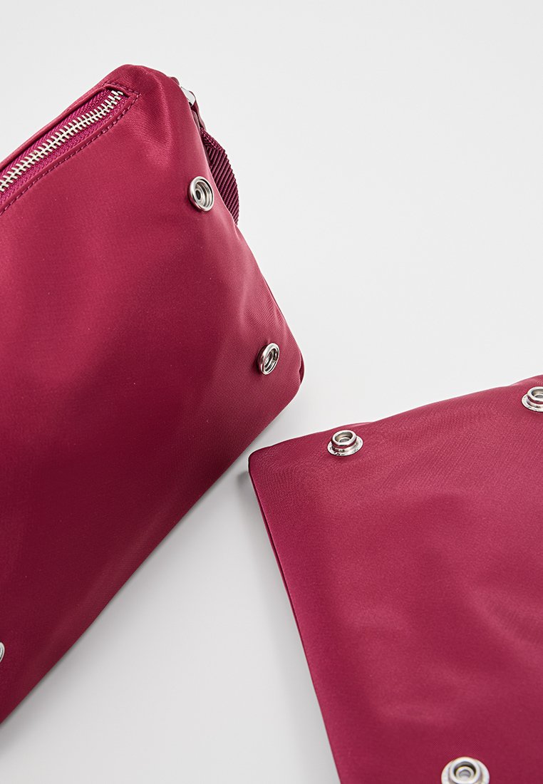 Bolsa de nailon burdeos con cierre de cremallera. Presenta detalles de botón de metal y una textura suave. Forma compacta y rectangular.
