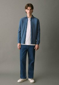 Giovane uomo in piedi contro un muro grigio, che indossa una camicia blu button-down sopra una maglietta bianca, jeans blu e sneakers bianche.