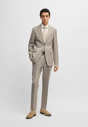Completo beige chiaro con giacca aderente e pantaloni, caratterizzato da una leggera texture. Abbinato a una camicia bianca e una cravatta beige, e scarpe eleganti color marrone chiaro.