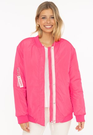 Junge Frau lächelt, trägt eine knallpinkfarbene Bomberjacke mit weißem Hemd und hellen Hosen vor weißem Hintergrund.