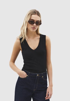 Kobieta w okularach przeciwsłonecznych w szylkretowych oprawkach, czarnej koszulce bez rękawów z dekoltem w szpic i ciemnoniebieskich dżinsach, stoi z jedną ręką w kieszeni.