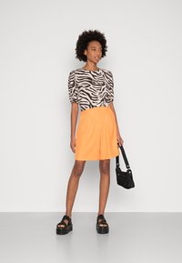 Blusa de manga curta com padrão de zebra em tons neutros, combinada com calções laranja de cintura alta até ao joelho e sandálias de plataforma pretas robustas.