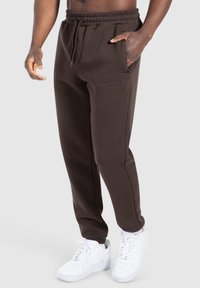 Mann in dunkelbraunen Jogginghosen mit elastischem Bund und weißen Sneakers, Hand in der Tasche, vor einem schlichten grauen Hintergrund.