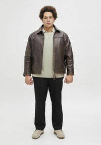 Veste en cuir marron avec un pantalon noir, chemise polo vert clair en dessous, et chaussures beiges. Le modèle se tient devant un fond uni.