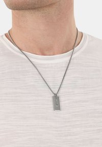 Collana con pendente in argento su una catena, caratterizzata da un'etichetta rettangolare con una superficie testurizzata e un logo impresso, indossata sopra una camicia bianca.