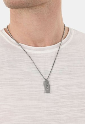 Collar de pendant de plata sobre una cadena, con una etiqueta rectangular de superficie texturizada y logo en relieve, usado sobre una camisa blanca.