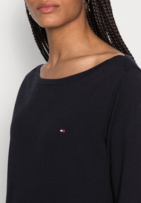 Marinblå stickad tröja med bred halsringning och korta ärmar. Har en liten logotyp med röda, vita och blå inslag på bröstet.