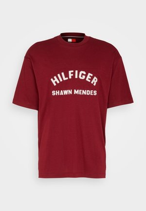Mørkerød bomulds T-shirt med korte ærmer, rund halsudskæring og hvid tekst, der står "HILFIGER SHAWN MENDES" i midten foran i en buet stil.