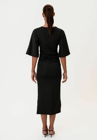 Schwarzes Kleid mit kurzen Glockenärmeln und einer taillierten Silhouette. Der Stoff ist glatt mit einer Raffung auf der Rückseite, die bis zur Mitte der Waden reicht.