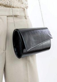 Bolso de mano negro de cuero con un diseño plegable, que cuenta con una correa de cadena y una textura suave, exhibido junto a unos pantalones beige.