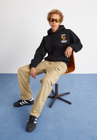 Felpa nera con grafica, pantaloni cargo beige e sneakers neri con strisce bianche. Il modello è seduto su una sedia di legno contro uno sfondo grigio chiaro.