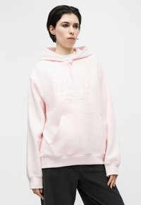 Νεαρό άτομο με κοντά σκούρα μαλλιά που φοράει ροζ ανοιχτό φούτερ με κουκούλα με την επιγραφή "New Balance Athletics" και μαύρο παντελόνι, στέκεται μπροστά σε λευκό φόντο.