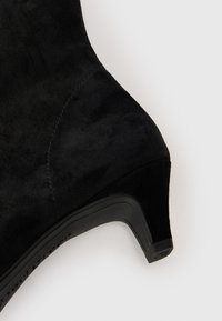 Botte de cheville en suede noir avec un talon fuselé, présentant une texture lisse, des coutures discrètes et une légère courbure au niveau de la cheville.
