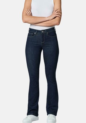 Person trägt dunkelblaue Bootcut-Jeans, weißes ärmelloses Oberteil und weiße Sneaker, steht mit verschränkten Armen vor weißem Hintergrund.