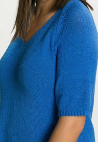 Modrý pletený svetr s výstřihem do V, krátkými rukávy a žebrovanými manžetami. Textura je hladká a mírně volnější.