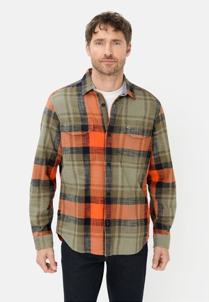 Man wearing green, orange, and black plaid button-up shirt over white undershirt, standing against plain white background.