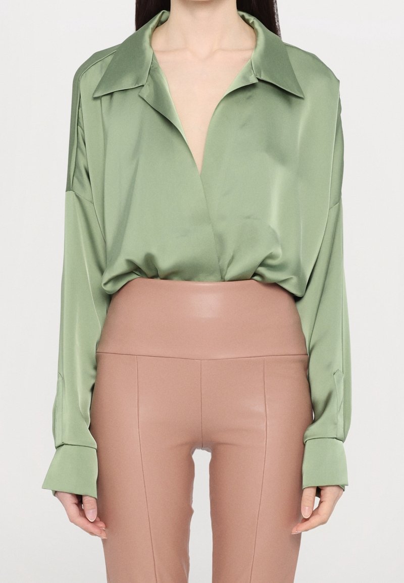Femme portant une chemise ample en satin vert clair rentrée dans un pantalon en cuir beige taille haute et ajusté sur un fond uni.