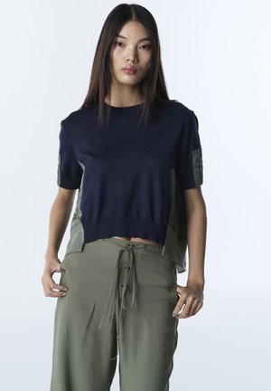 Joven mujer con una camiseta de manga corta azul marino y pantalones sueltos verde oliva con cordones, de pie contra un fondo liso y claro.