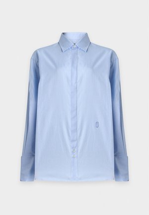 Femme portant une chemise ample bleu clair boutonnée, une courte jupe en jean avec des patchs, et des chaussures plates pointues noires avec des boucles décoratives.