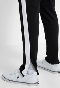 Person med svarta byxor med vita sidosträck och dragkedjor vid anklarna, tillsammans med vita låga sneakers med Polo-logotyp.