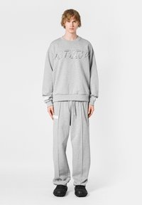 Sudadera gris con un logo, combinada con pantalones de chándal grises que tienen bolsillos laterales y acentos de cadena. El modelo lleva calzado negro.