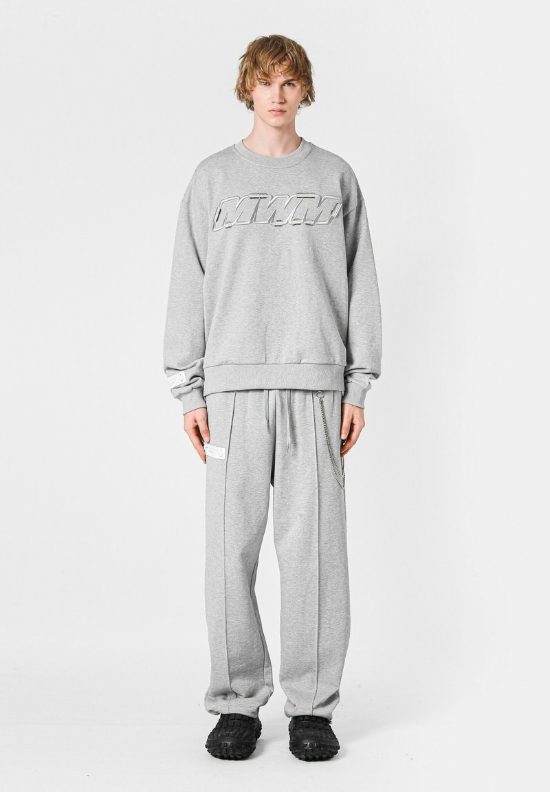 Sudadera gris con un logo, combinada con pantalones de chándal grises que tienen bolsillos laterales y acentos de cadena. El modelo lleva calzado negro.