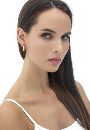 Goldene Creolen schmücken das Ohr eines Models, mit einer glatten, polierten Oberfläche. Sie hat lange, glatte braune Haare und einen neutralen Hautton.
