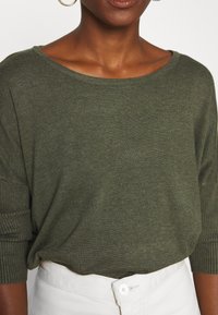 Olivgrön, löst sittande stickad tröja med vid halsringning och ribbade ärmslut, i kombination med vita högmidjade byxor.