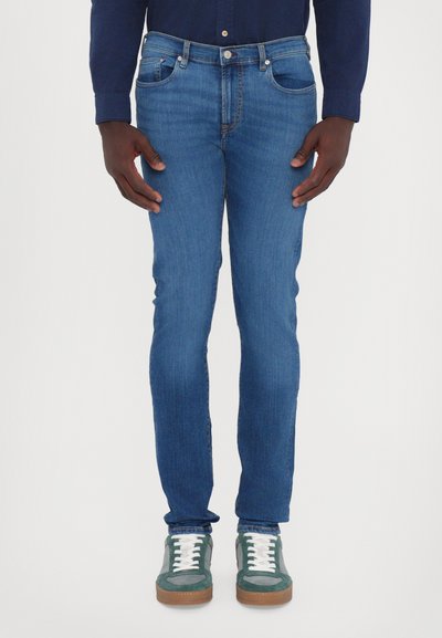 Slim-fit modré džíny s jemným vyblednutím, pěti kapsami a klasickým zapínáním na knoflík, doplněné zeleno-bílými teniskami.