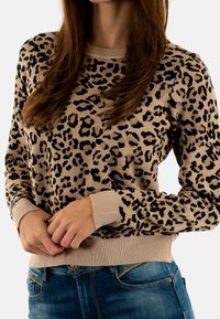 Mujer con un suéter de estampado de leopardo en tonos marrón y negro, y Jeans azules, con las manos entrelazadas de forma relajada a la altura de la cintura, recortado por debajo de la barbilla.