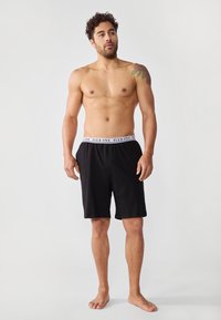 Sorte athletic shorts med elastisk midjebånd som har en kontrasterende logo. Stoffet virker glatt, og passformen er løs.