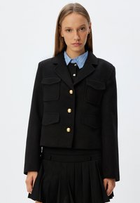 Zwarte cropped jas met gouden knopen, voorzien van twee voorzakken. Gekocht over een zwart top en een blauw gestreept overhemd met kraag.