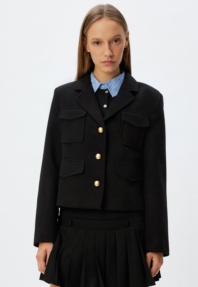 Zwarte cropped jas met gouden knopen, voorzien van twee voorzakken. Gekocht over een zwart top en een blauw gestreept overhemd met kraag.