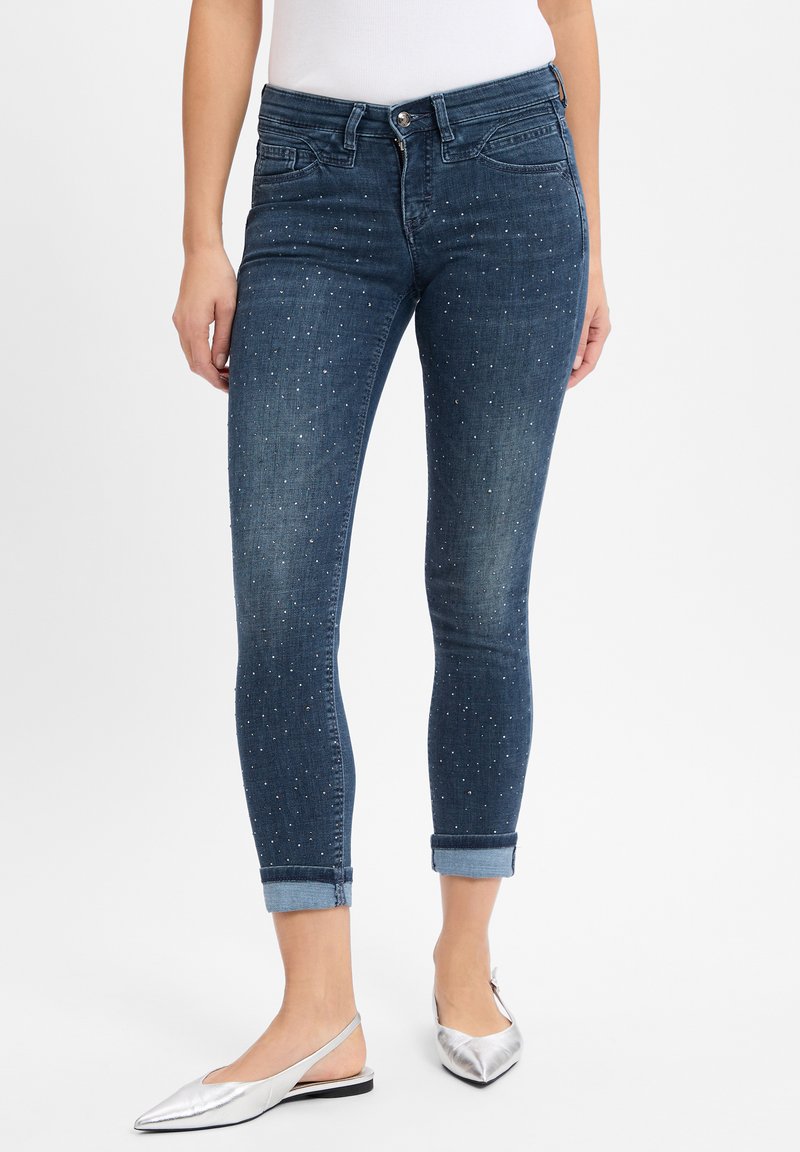 Dunkelblaue Skinny-Jeans mit umgekrempelten Säumen, verziert mit kleinen silbernen Nieten, im klassischen Fünf-Taschen-Design und mit vorderem Knopfverschluss.